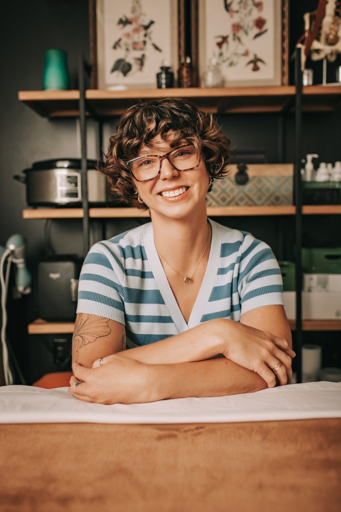 Jenny Gabris smiling in a treatment room, wearing glasses and a blue-and-white striped top.