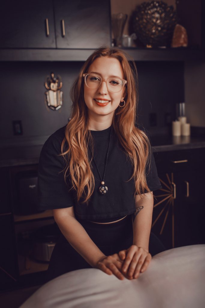 Joanna, a Precision Wellness Esthetician, smiling in a treatment room with dark cabinetry, wearing glasses and a black top.