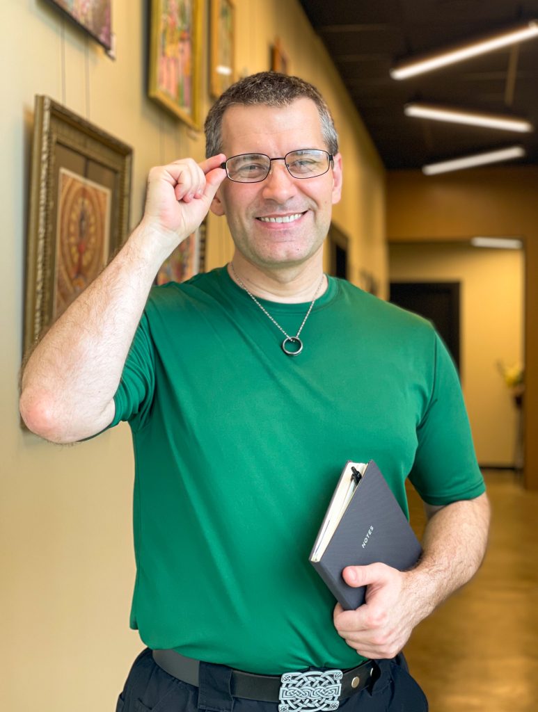 Chad Bright, Licensed Massage Therapist, smiling in a hallway wearing glasses and a green shirt, holding a notebook.