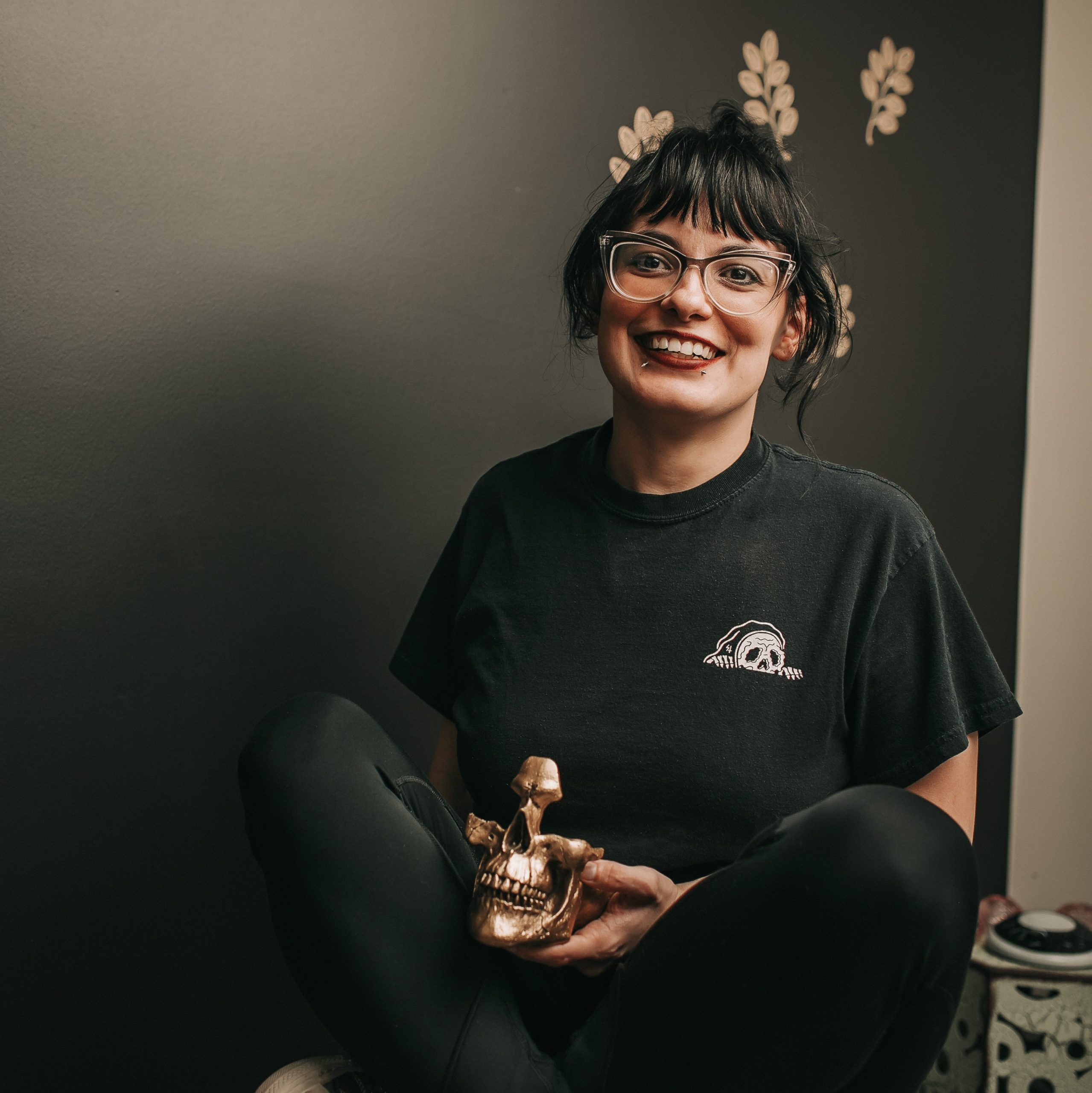 Kaya, LMT, smiling while seated against a dark wall, wearing glasses and a black top, holding a small gold skull decor piece.