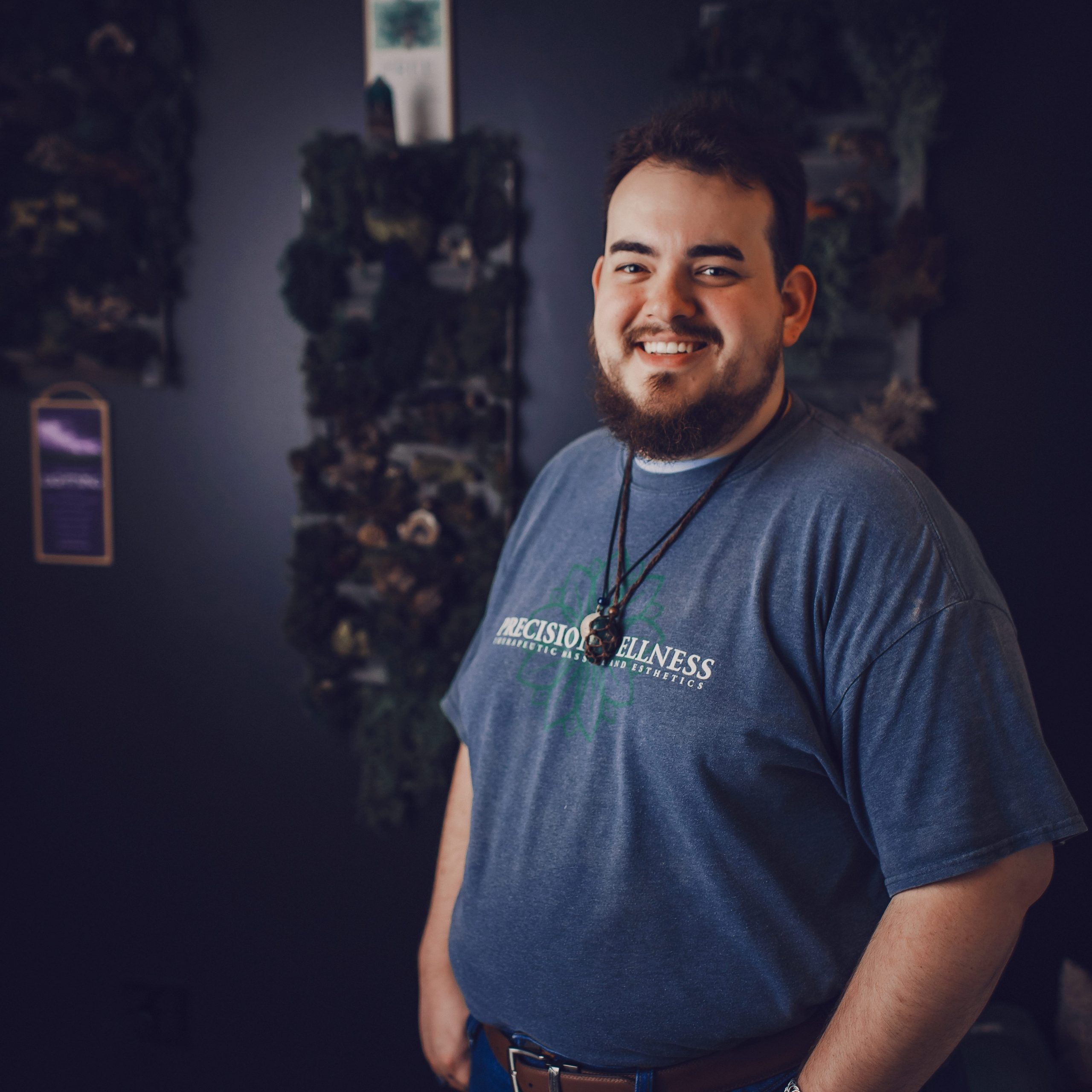 Grant, LMT Lead, smiling in a treatment room wearing a Precision Wellness t-shirt, with greenery wall decor in the background.