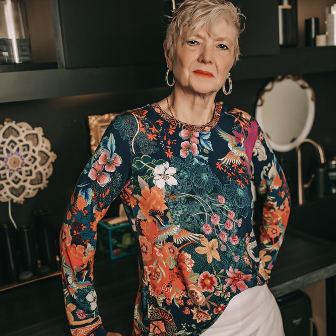 Jacqui, LMT, standing in a treatment room wearing a colorful floral top and white pants, with dark cabinetry in the background.