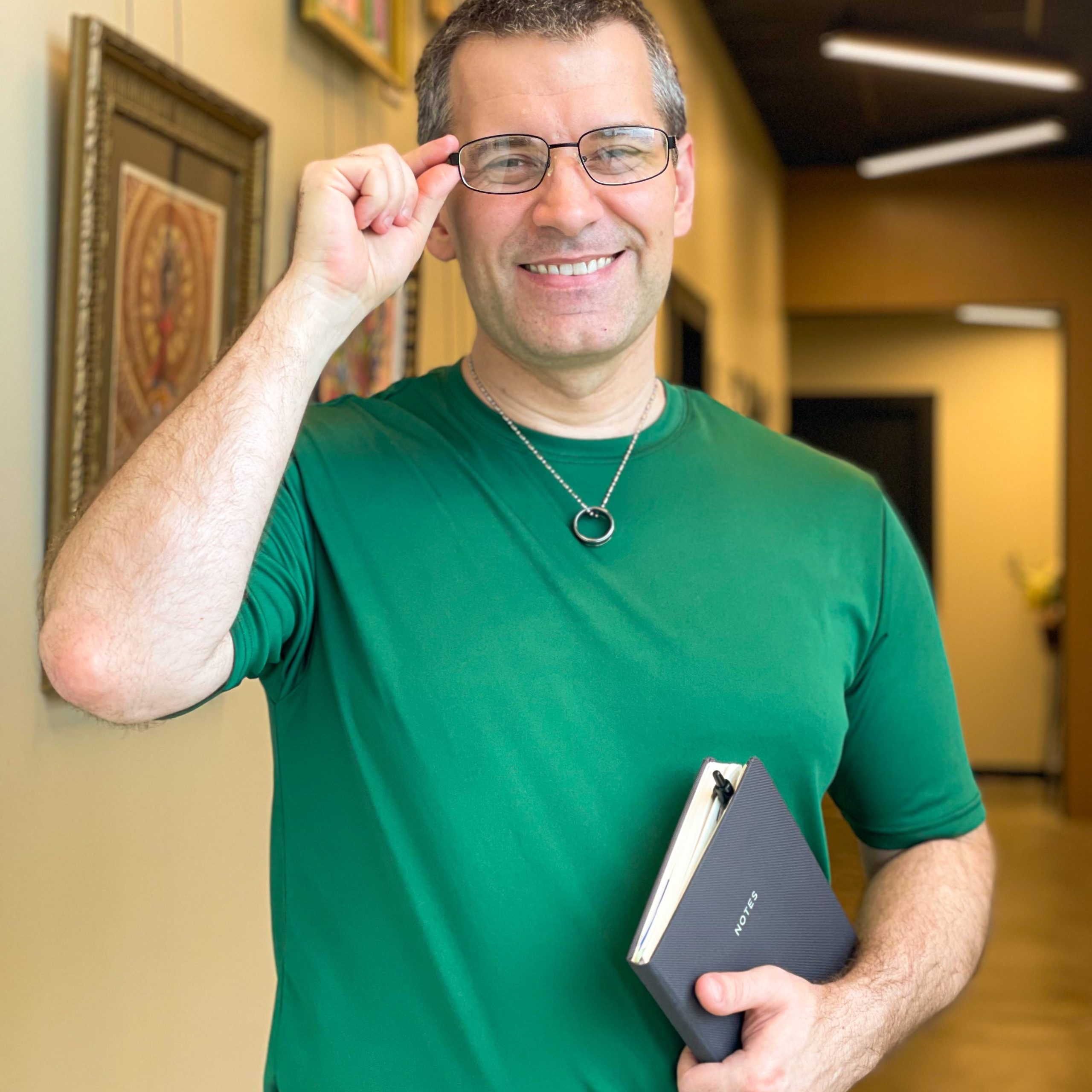 Chad Bright, Licensed Massage Therapist, smiling in a hallway wearing glasses and a green shirt, holding a notebook.