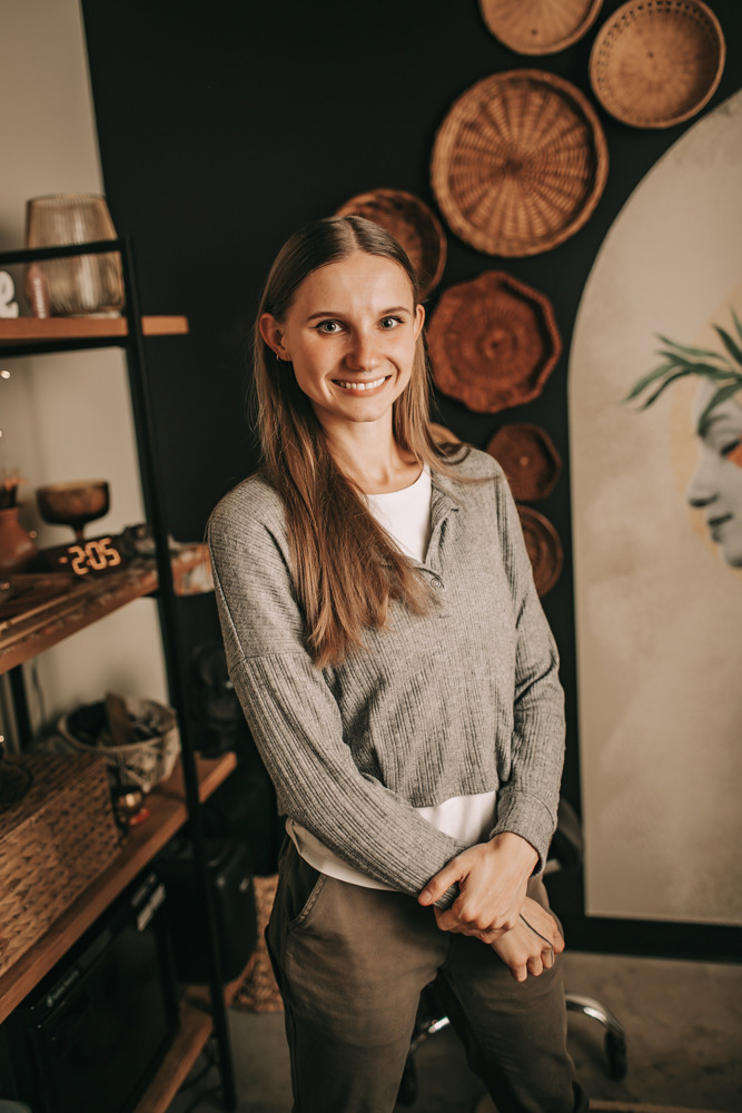 Olivia, LMT, smiling in a treatment room wearing a gray top, with woven wall decor and spa shelving in the background.