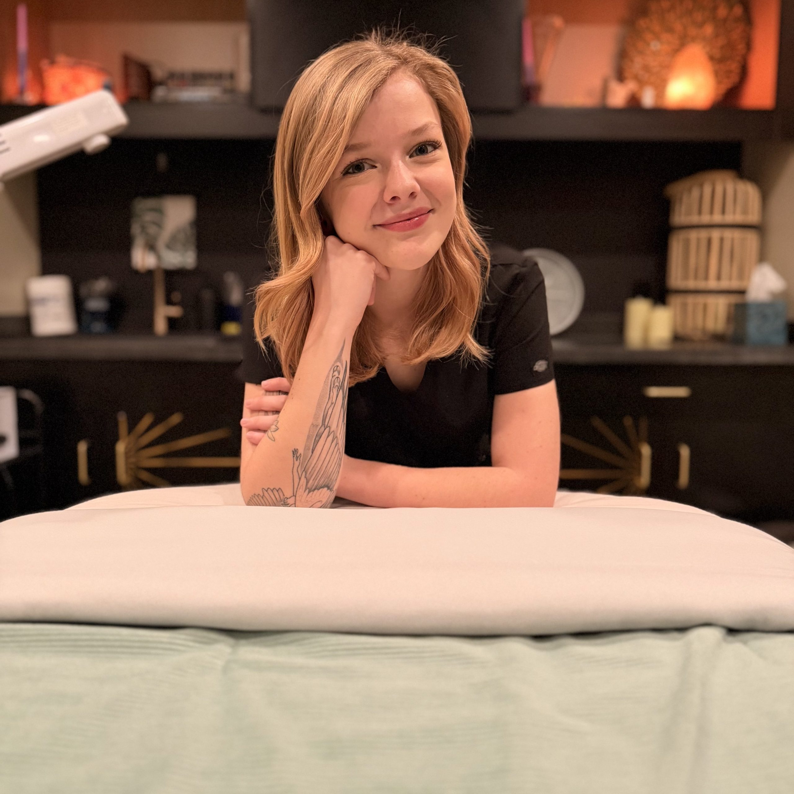 Sarah Ritter leaning on a treatment table in a spa room, smiling, with warm lighting and shelves in the background.