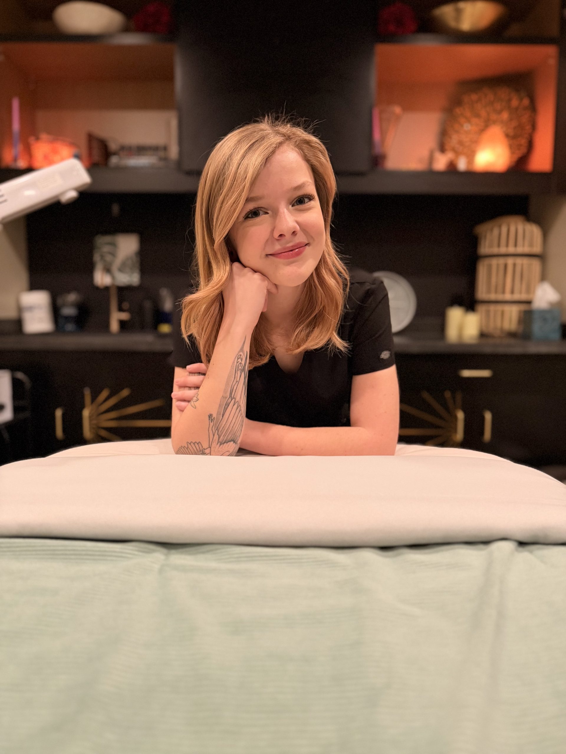 Sarah Ricketts leaning on a treatment table in a spa room, smiling, with warm lighting and shelves in the background.