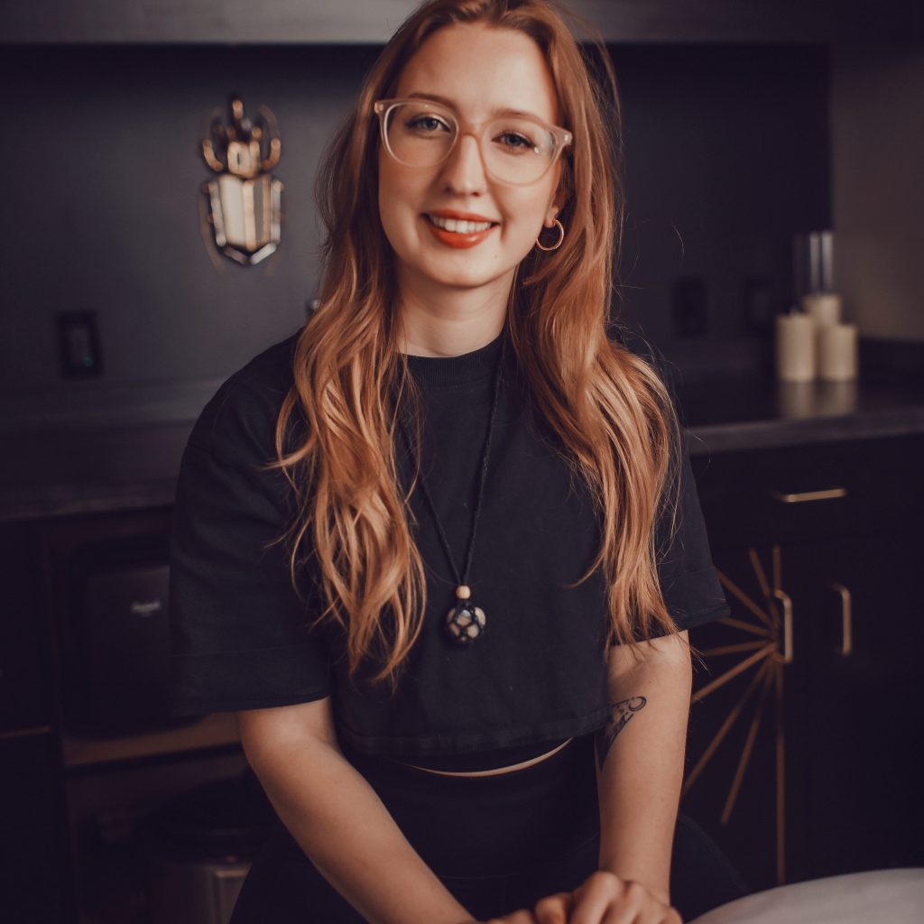 Joanna, a Precision Wellness Esthetician, smiling in a treatment room with dark cabinetry, wearing glasses and a black top.