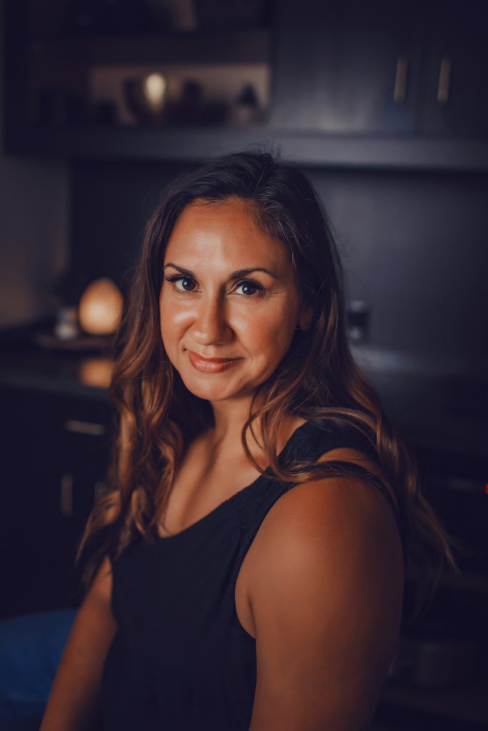 Serrenia, Lead Esthetician, smiling in a treatment room with warm lighting and dark cabinetry in the background.