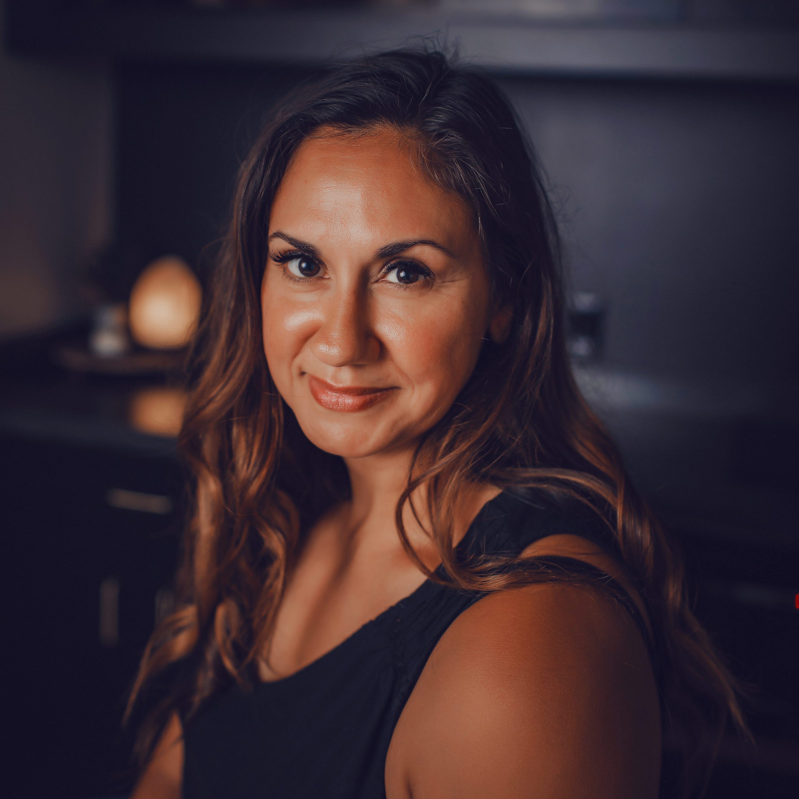 Serrenia, Lead Esthetician, smiling in a treatment room with warm lighting and dark cabinetry in the background.