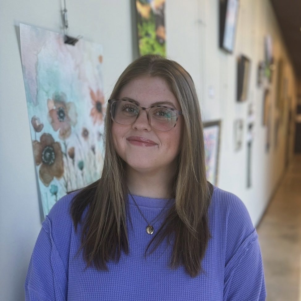 Haley LaCraze smiling in a bright hallway at Precision Wellness, wearing glasses and a purple top with artwork in the background.