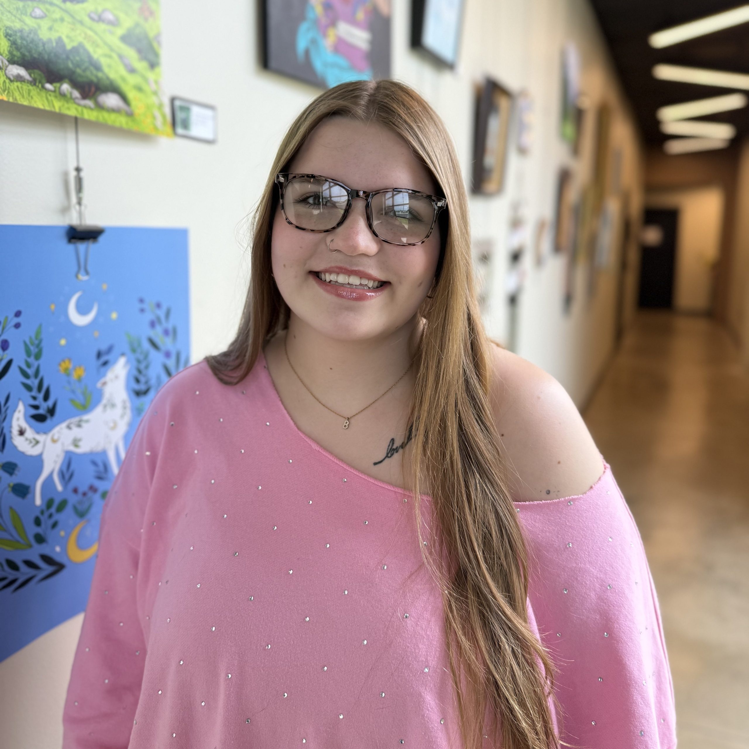 Selena Brown smiling in a bright hallway at Precision Wellness, wearing glasses and a pink top with artwork in the background.